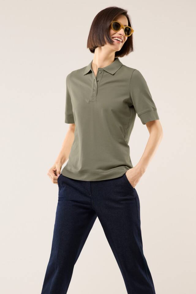 Eine Frau steht vor hellem Hintergrund und trägt ein kurzärmeliges, olivgrünes Poloshirt sowie eine dunkelblaue Hose. Sie lächelt und hat die Hände lässig in den Hosentaschen.
