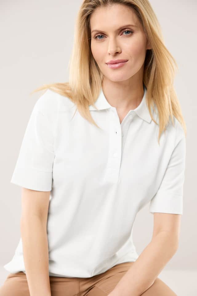 Eine Frau trägt ein weißes, kurzärmeliges Poloshirt und sitzt leicht nach vorne gelehnt vor einem neutralen Hintergrund. Sie blickt freundlich in die Kamera.