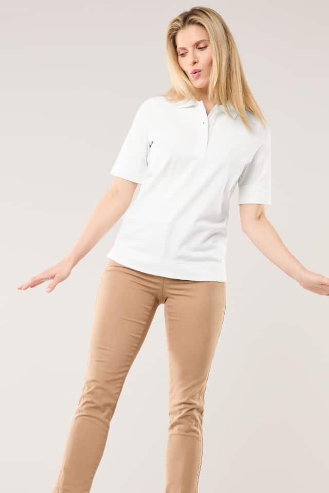 Eine Frau steht vor neutralem Hintergrund und trägt ein weißes Poloshirt mit kurzen Ärmeln sowie eine beigefarbene, schmal geschnittene Hose. Sie hält die Arme leicht zur Seite ausgestreckt.