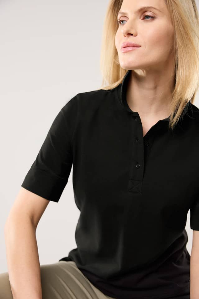 Eine Frau trägt ein schwarzes Poloshirt mit kurzen Ärmeln und sitzt vor einem hellen Hintergrund. Die Aufnahme zeigt Oberkörper und Gesicht in einer ruhigen Pose.