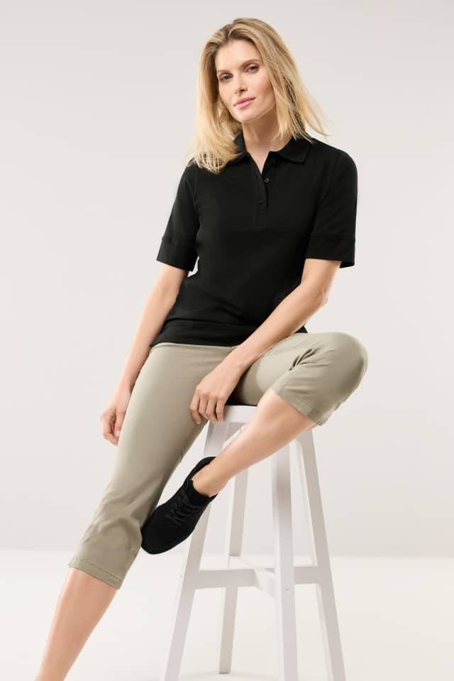 Eine Frau sitzt auf einem weißen Hocker und trägt ein schwarzes Poloshirt mit kurzen Ärmeln sowie eine beigefarbene, knielange Hose. Sie blickt leicht lächelnd in die Kamera.
