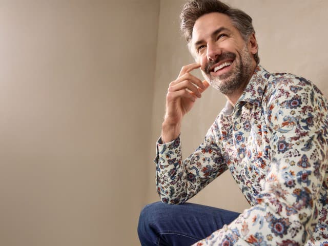 Eine Person sitzt lächelnd und trägt ein helles Hemd mit buntem Blumenmuster, kombiniert mit einer blauen Jeans