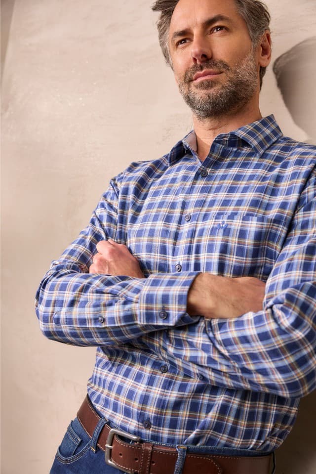 Ein Mann mit grauem Haar und Bart trägt ein blau kariertes Hemd mit Brusttasche, eine dunkelblaue Jeans und einen braunen Gürtel und steht mit verschränkten Armen vor einer hellen Wand.