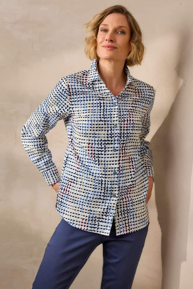 Eine Frau mit blonden, welligen Haaren trägt ein langärmliges, gemustertes Hemd in Blau-, Weiß- und Rottönen sowie eine dunkelblaue Hose und steht mit den Händen in den Taschen vor einer hellen Wand.