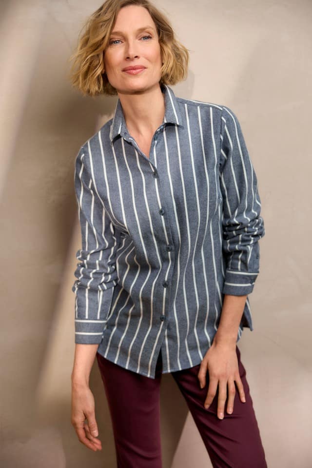 Eine Frau mit blondem, welligem Haar trägt ein blau-grau gestreiftes Hemd mit weißen Längsstreifen und eine weinrote Hose und steht leicht nach vorn gelehnt vor einer hellen Wand.