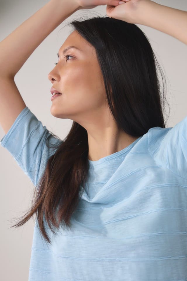 Nahaufnahme einer Frau mit langen dunklen Haaren in einem hellblauen T-Shirt, die beide Arme über dem Kopf verschränkt und nach links blickt.
