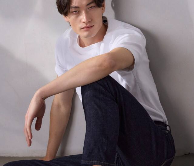 Ein Mann sitzt am Boden vor einer hellen Wand und trägt ein weißes T‑Shirt, dunkle Jeans und weiße Sneaker.