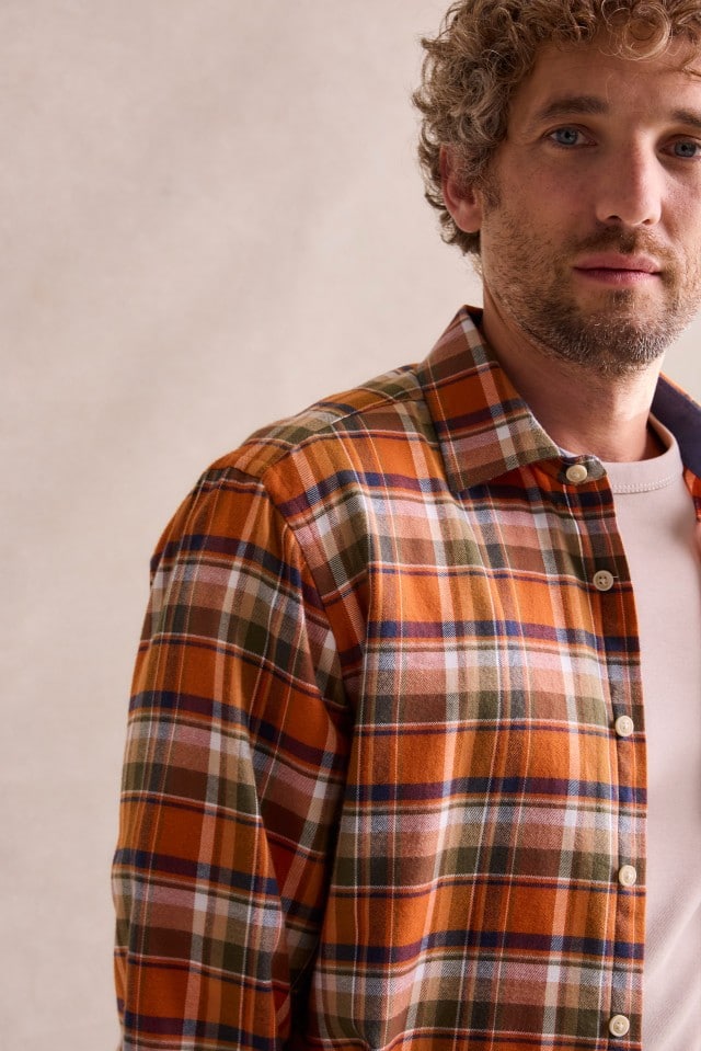 Ein Mann mit lockigem Haar trägt ein orange-braun kariertes Hemd über einem hellen T-Shirt und blickt ruhig in die Kamera.