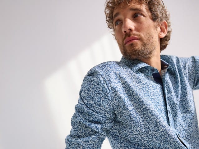 Ein Mann trägt ein blaues, floral gemustertes Hemd und blickt seitlich nach oben.