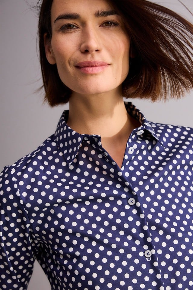 Eine Person mit kinnlangem Haar trägt eine dunkelblaue Bluse mit weißen Punkten und lächelt leicht in die Kamera.