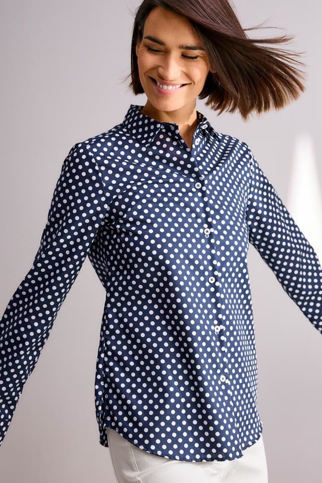 Eine Person mit kinnlangem Haar trägt eine dunkelblaue Bluse mit weißen Punkten und dreht sich lächelnd zur Seite.