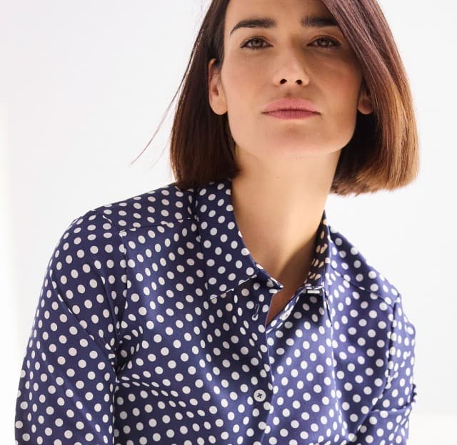 Eine Person mit kinnlangem Haar trägt eine dunkelblaue Bluse mit weißen Punkten und blickt leicht zur Seite.