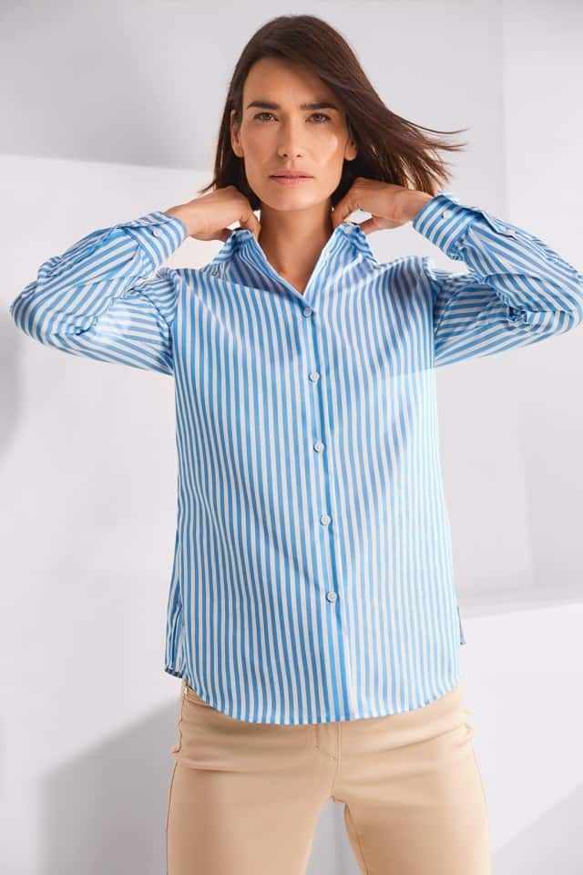 Eine Person mit schulterlangem Haar trägt ein blau‑weiß gestreiftes Hemd und richtet den Kragen mit beiden Händen.