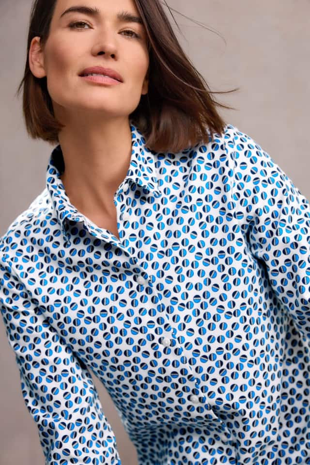 Eine Frau mit schulterlangem, braunem Haar trägt eine weiße Bluse mit einem blauen, runden Muster und blickt leicht lächelnd nach vorn.