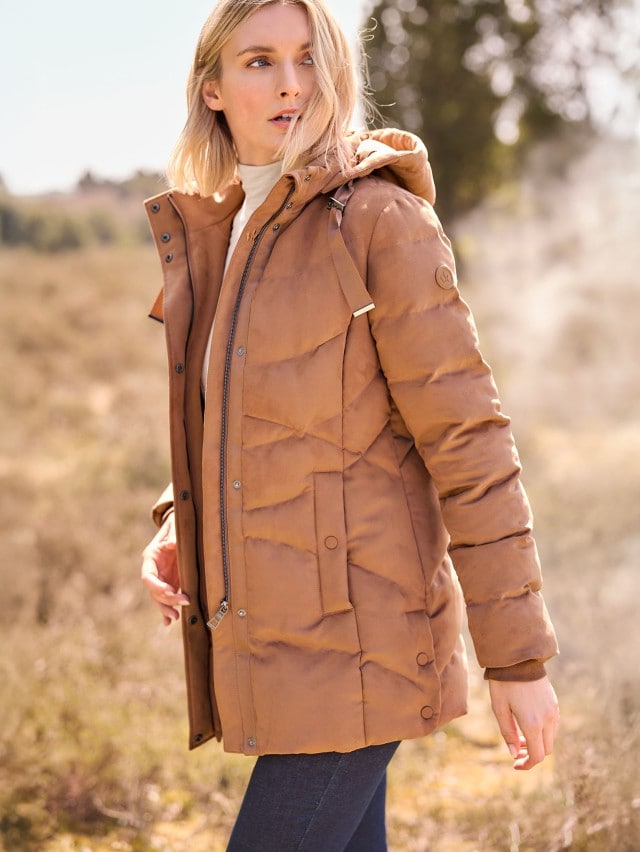 Eine Person steht im Freien und trägt eine hellbraune, wattierte Winterjacke mit Kapuze über einem beigen Rollkragenpullover und einer dunklen Hose.