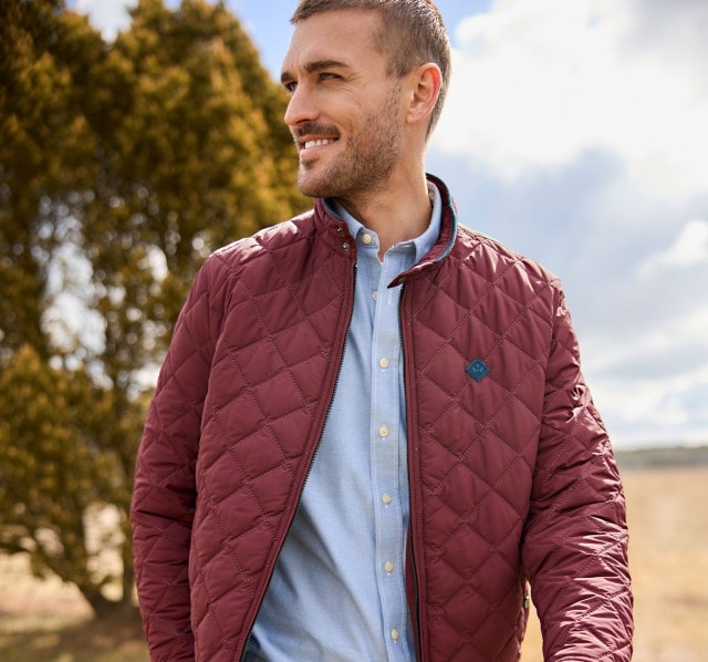 Eine Person steht im Freien und trägt eine weinrote, gesteppte Jacke über einem hellblauen Hemd und einer grauen, fein karierten Hose.
