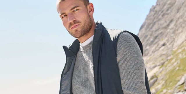 Ein Mann gesteht vor einem Gebirge und trägt eine Weste in Marine, darunter einen grauen Pullover.