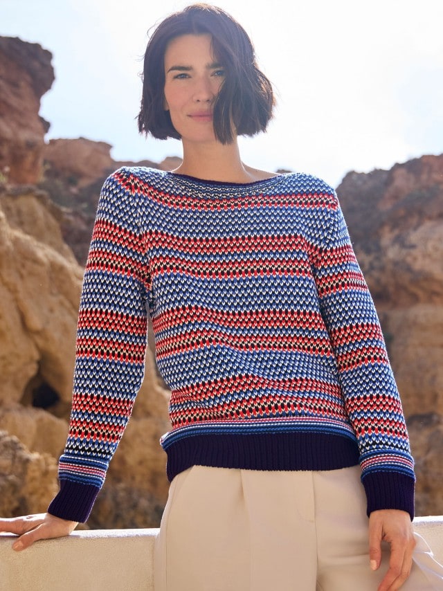 Frau in einem rot-blau-weiß-gestreiften Strickpullover und heller Hose steht an einer Mauer vor Felsen und schaut frontal in die Kamera.
