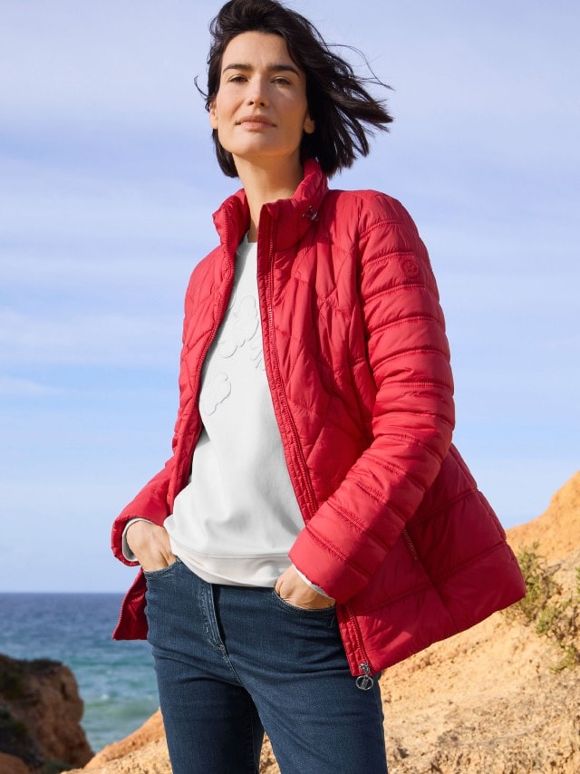 Frau in einer roten Steppjacke über einem weißen Pullover und Jeans steht auf einem Felsen am Meer und schaut in die Kamera.