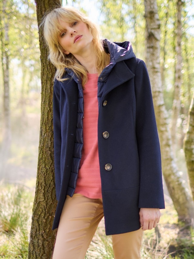 Eine Frau trägt einen dunkelblauen Mantel mit großen Knöpfen und gesteppter Kapuze über einem lachsfarbenen Pullover, kombiniert mit einer beigefarbenen Hose im lässig-eleganten Stil.