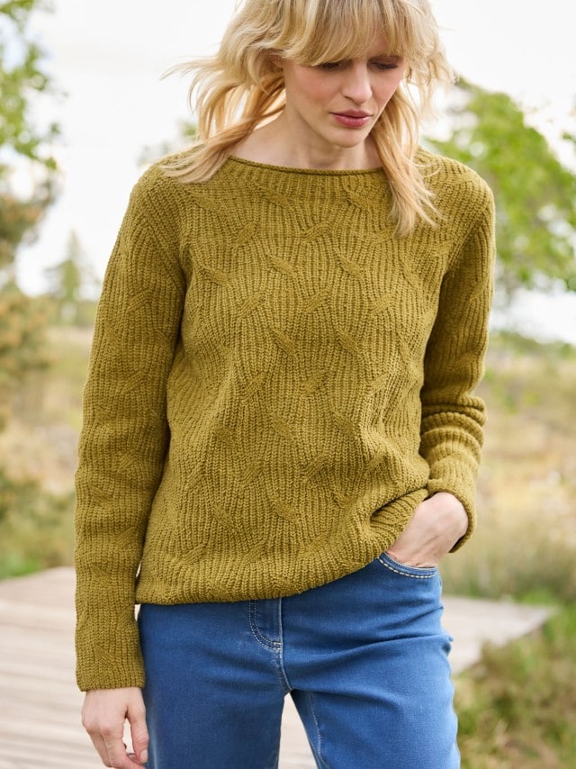 Eine Frau trägt einen senfgelben Strickpullover mit plastischem Strukturmuster, kombiniert mit einer blauen Jeans im legeren, modernen Stil.