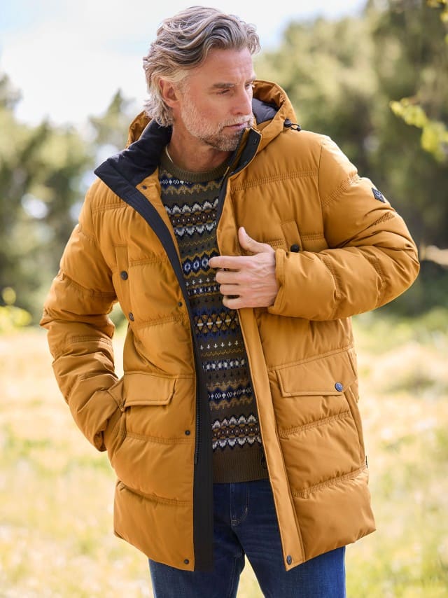 Ein Mann trägt eine senfgelbe Steppjacke über einem Strickpullover mit Norwegermuster und Jeans; der Stil wirkt warm, gemütlich und herbstlich.