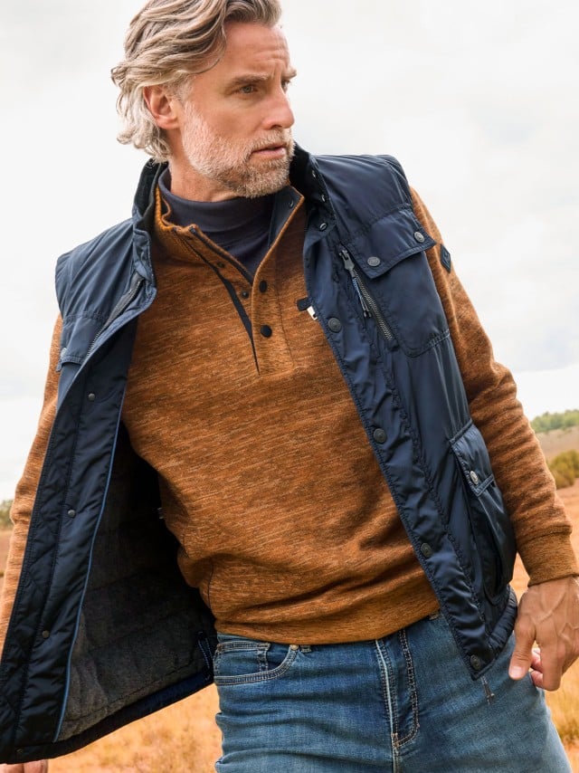 Ein Mann trägt einen rostbraunen Pullover mit Knopfleiste und eine dunkelblaue Steppweste zu Jeans; der Look wirkt lässig und herbstlich-sportlich.