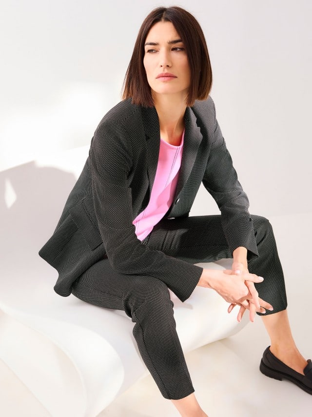 Eine Frau sitzt auf einem futuristischen Stuhl und trägt einen Reversblazer Dots in Schwarz/Weißt, dazu die passende Hose und ein Edeljerseyshirt mit Zierstich in Malve.


