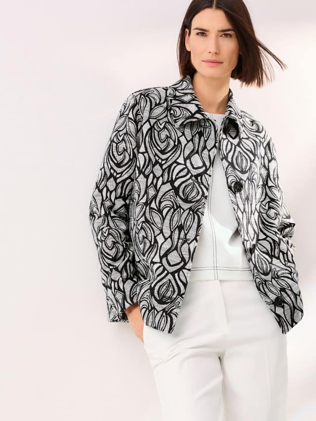 Eine Frau trägt eine schwarz/weiße Jacquard Jacke, darunter ein weißes Edeljerseyshirt mit Zierstich und dazu eine Marlenehose mit Struktur in Creme.