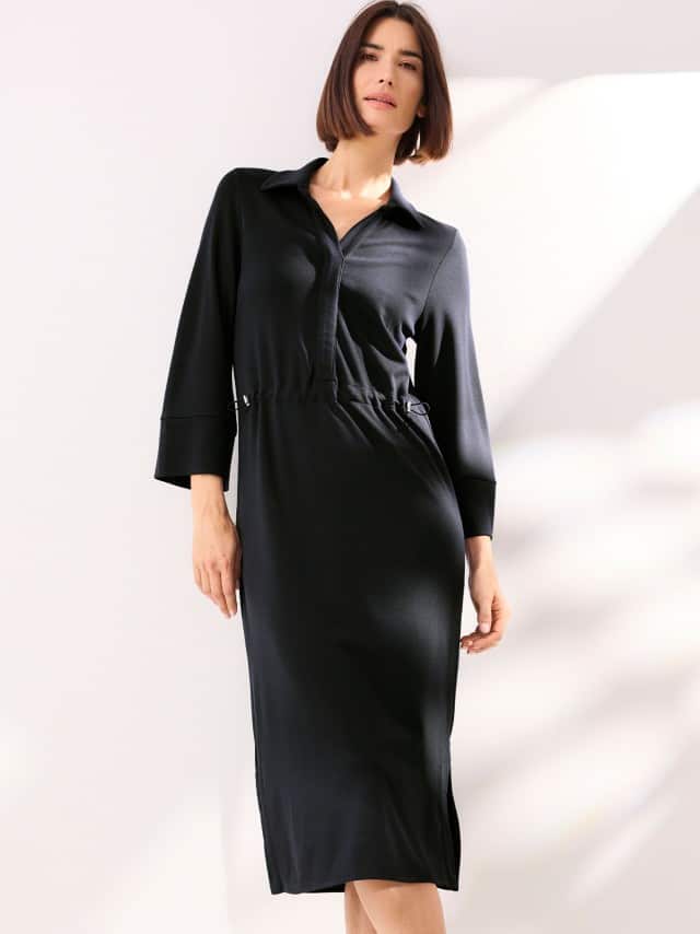 Eine Frau steht vor einer hellen Wand und trägt ein schwarzes Midi Jerseykleid.