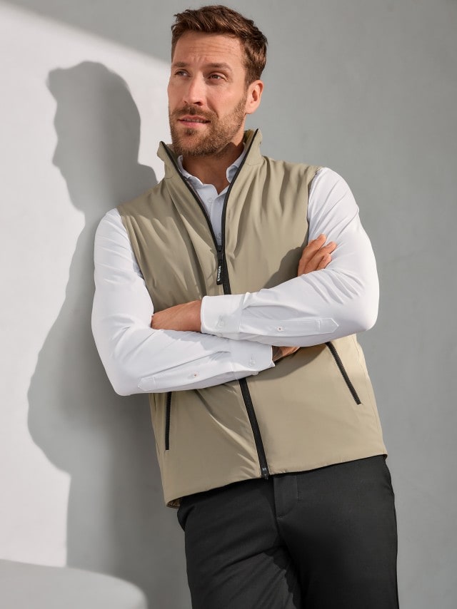 Ein Mann steht an einer Wand angelehnt und trägt ein weißes Hemde, darüber eine Daunen Hybrid Weste in Beige und dazu eine schwarze Hose. 