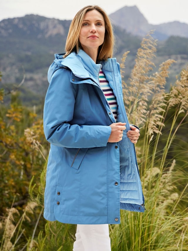 Frau in einer hellblauen Jacke und gestreiftem Oberteil steht in einer Wiese und schaut in die Ferne.
