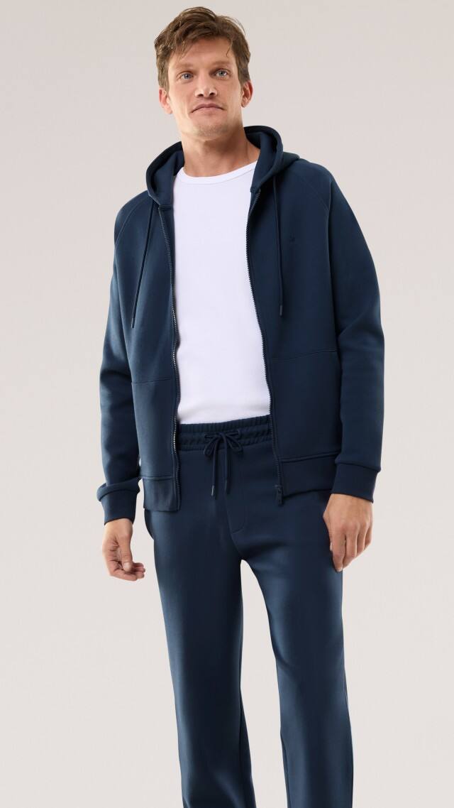 Ein Mann trägt einen dunkelblauen Freizeitanzug mit Hoodie und passender Hose, kombiniert mit einem schlichten weißen T‑Shirt. Der Look wirkt sportlich, modern und bequem.