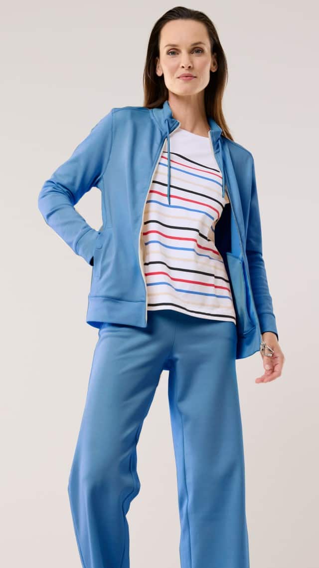 Eine Frau trägt einen blauen Freizeitanzug mit Jacke und weiter Hose, kombiniert mit einem hellen T‑Shirt mit bunten Streifen. Der Look wirkt sportlich und entspannt.