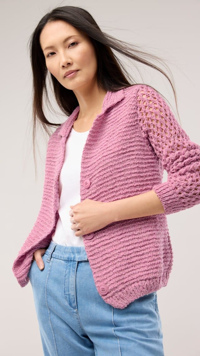 Eine Frau trägt eine rosafarbene Strickjacke mit durchbrochenen Ärmeln über einem weißen Oberteil und einer hellblauen Hose.
