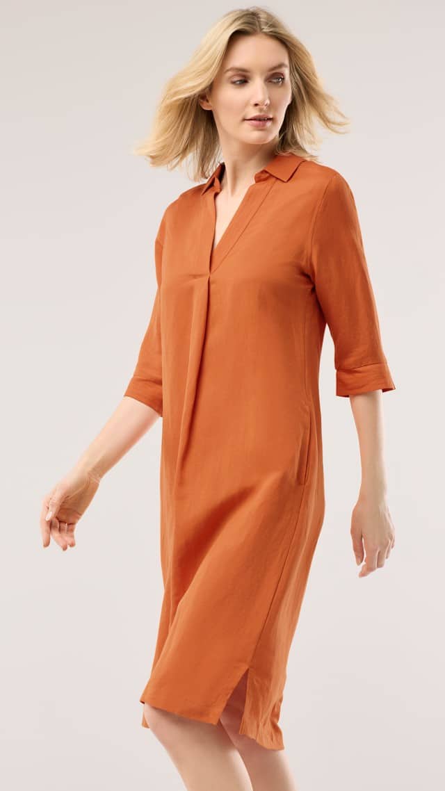 Eine Frau trägt ein knielanges, orangefarbenes Kleid mit halblangen Ärmeln und läuft leicht seitwärts.