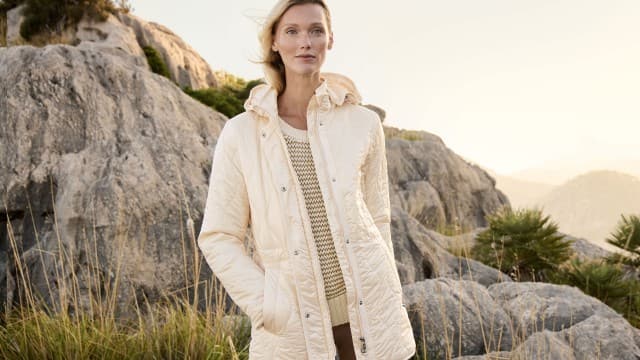 Eine Frau steht vor einer felsigen Landschaft und trägt eine helle Jacke über einem gestrickten Oberteil. Eine Frau steht vor einer felsigen Landschaft und trägt eine helle Jacke über einem gestrickten Oberteil.