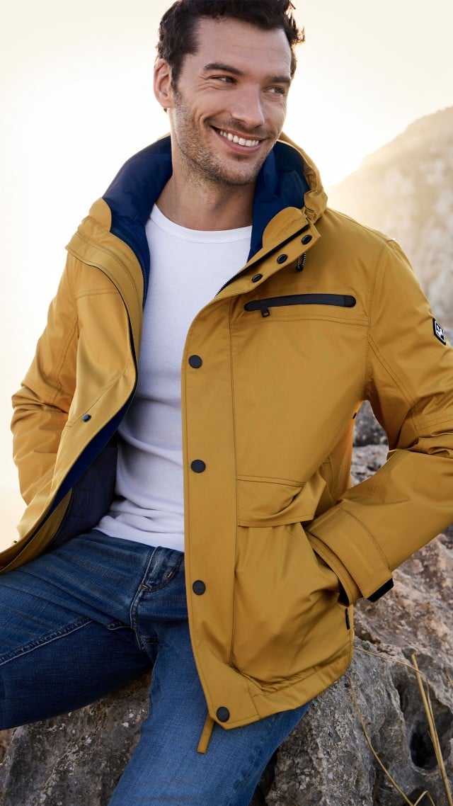 Ein Mann sitzt in einer Felslandschaft und trägt eine gelbe Jacke über einem weißen T‑Shirt und Jeans. Ein Mann sitzt in einer Felslandschaft und trägt eine gelbe Jacke über einem weißen T‑Shirt und Jeans.