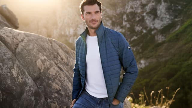 Ein Mann steht in einer Felsenlandschaft und trägt eine blaue Steppjacke über einem weißen T‑Shirt und Jeans. Ein Mann steht in einer Felsenlandschaft und trägt eine blaue Steppjacke über einem weißen T‑Shirt und Jeans.