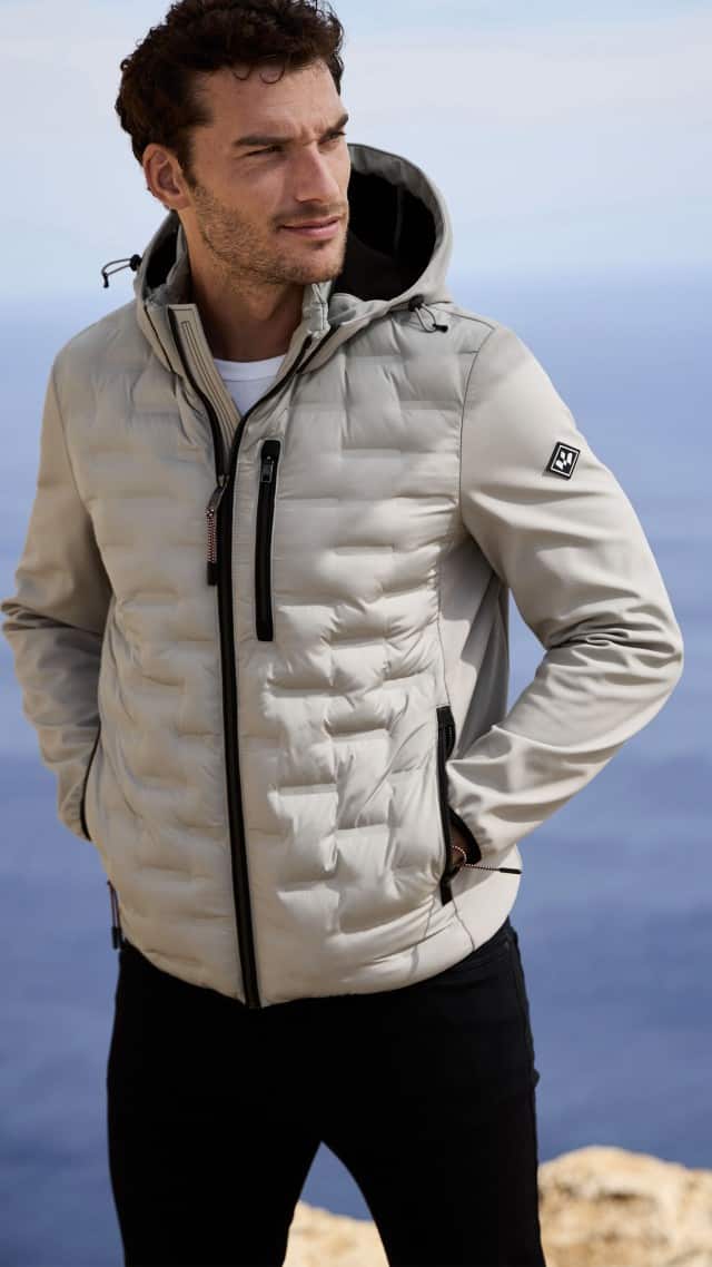 Ein Mann steht vor einem Meerblick und trägt eine hellgraue Kapuzenjacke mit schwarzen Reißverschlüssen. Ein Mann steht vor einem Meerblick und trägt eine hellgraue Kapuzenjacke mit schwarzen Reißverschlüssen.