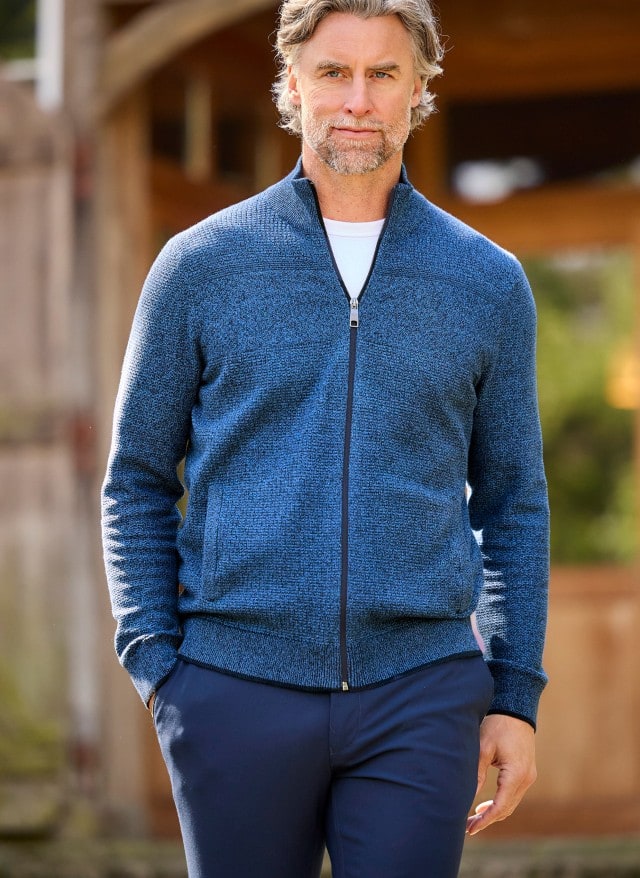 Ein Mann in einer blau-melierten Strickjacke und blauer Hose läuft locker vor eine Gebäude entlang und schaut zur Seite.