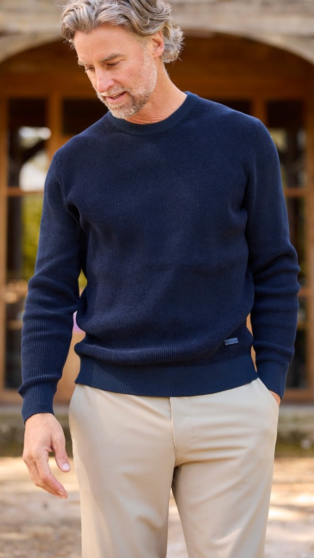 Ein Mann steht vor einem Gebäude, trägt einen dunkelbauen Ripppullover und eine helle Hose und schaut nach unten.