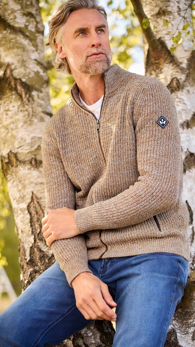 Ein Mann lehnt an einem Baum, trägt eine braun-melierte Strickjacke über einem weißen Shirt und Jeans und schaut zur Seite.