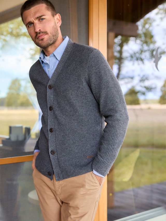 Ein Mann steht vor einer großen Fensterfront und trägt einen Cardigan, darunter ein hellblaues Hemd und dazu eine Hose in Beige. Ein Mann steht vor einer großen Fensterfront und trägt einen Cardigan, darunter ein hellblaues Hemd und dazu eine Hose in Beige.