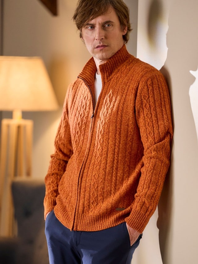 Ein Mann steht angelehnt an einer Wand und trägt eine Cashmino Zopf-Strickjacke in Terra, darunter ein weißes Shirt und dazu eine Jeans. Ein Mann steht angelehnt an einer Wand und trägt eine Cashmino Zopf-Strickjacke in Terra, darunter ein weißes Shirt und dazu eine Jeans.