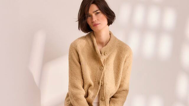 Eine Frau vor einem neutralen Hintergrund trägt die Baby Alpaka Strickjacke in der Farbe Sand.