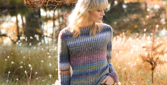 Ein blonde Frau steht auf einer herbstlichen Wiese, schaut zur Seite und trägt einen Grobstrick-Pullover mit Farbverlauf. Ein blonde Frau steht auf einer herbstlichen Wiese, schaut zur Seite und trägt einen Grobstrick-Pullover mit Farbverlauf.