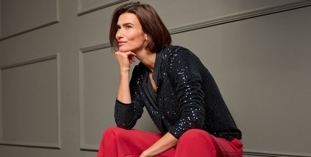 Eine Frau sitzt auf einem Stuhl, ist nach vorne gelehnt und trägt eine schwarze Paillettenjacke, dazu eine rote Hose. Eine Frau sitzt auf einem Stuhl, ist nach vorne gelehnt und trägt eine schwarze Paillettenjacke, dazu eine rote Hose.