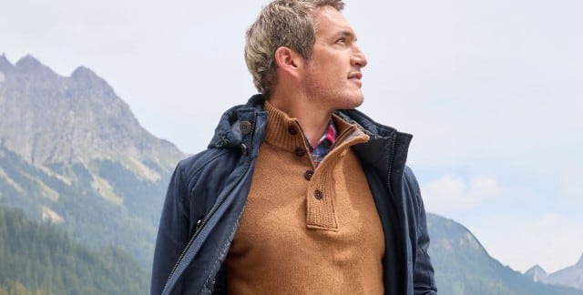 Ein Mann steht vor einem Gebirge, schaut zur Seite und trägt den Ultraskin Leichtparka in Marine, darunter einen Premium Lammwoll-Troyer in Camel.