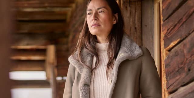 Eine Frau steht vor einer Hütte und trägt einen Rollkragen-Pullover, darüber den Premium Lammfell Wendemantel in Taupe.